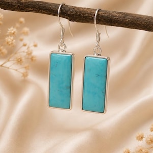 Puede incluir: Un par de pendientes colgantes con piedras turquesa rectangulares engastadas en marcos de metal plateado pulido. Cada pendiente cuelga de un sencillo gancho plateado, mostrando el vibrante color azul verdoso y las vetas naturales.