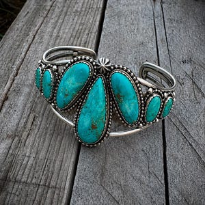 May include: A silver cuff bracelet featuring seven teardrop-shaped turquoise stones arranged in a cluster. The central stone is the largest, flanked by three smaller stones on each side. Each stone is set in a beaded silver frame with a star detail.