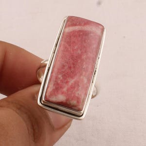 Peut inclure: Une bague rectangulaire ornée d'une pierre précieuse de rhodochrosite rose et blanche polie, sertie dans une monture en argent lisse. La pierre présente une texture marbrée naturelle avec des nuances de rose et de blanc, encadrée par un anneau.