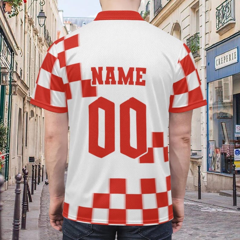 Op de afbeelding: Wit en rood geblokt sportshirt met het woord "NAME" en het nummer "00" in rood. Het shirt heeft een rode kraag en mouwen. De achtergrond is een Europese straatsc&egrave;ne.