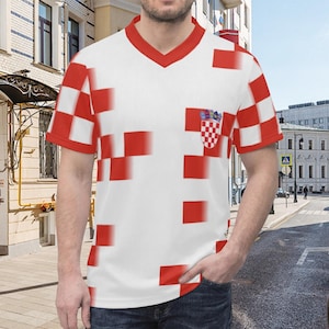 Op de afbeelding: Wit V-hals T-shirt met rood geblokt patroon en rode afwerking. Voorzien van een Kroatisch wapen op de borst. Het ontwerp van het shirt bevat de kleuren van de Kroatische vlag.