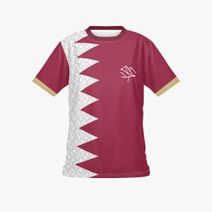 May include: Maroon t-shirt with a white and grey patterned design on the left side and a jagged edge design. The shirt has a white emblem on the right chest and gold trim on the sleeves.