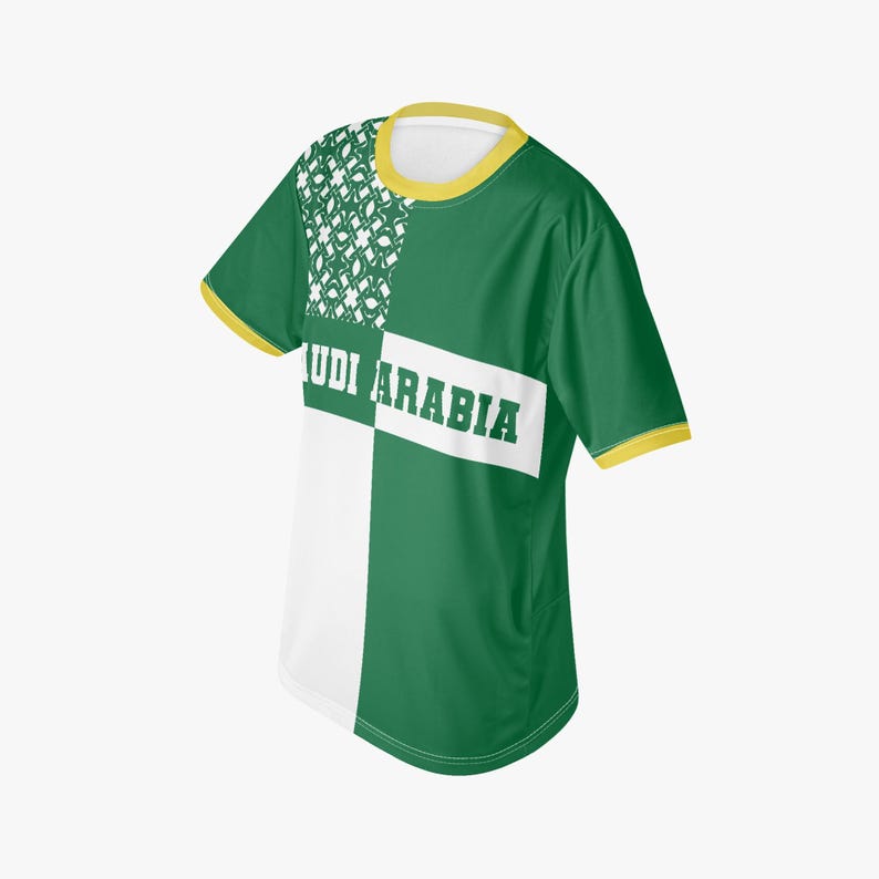 以下が含まれることがあります： 「SAUDI ARABIA」の白文字が入った緑と白のTシャツ。黄色の襟と袖口のトリムが付いています。デザインは、左上に白い模様が入った緑と白のカラーブロックパターンが特徴です。