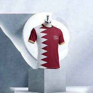 May include: A maroon t-shirt with a white and grey zigzag pattern on the left shoulder and a small white emblem on the right chest. The sleeves and collar have gold trim. The shirt is displayed on a mannequin.