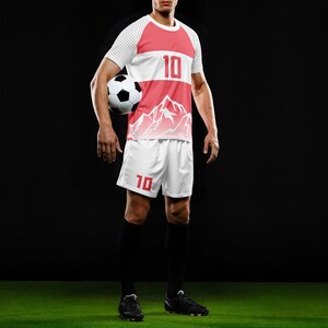 May include: A soccer uniform set in red and white, featuring a jersey and shorts. The jersey has a mountain graphic and the number 10. The shorts also display the number 10. The player is holding a soccer ball.