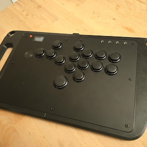 Puede incluir: Mando de lucha arcade negro con un asa y múltiples botones negros. El controlador tiene una pequeña pantalla y varias luces indicadoras. Los botones están dispuestos en un diseño estándar para juegos de lucha.