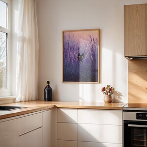 May include: A framed print depicting a black cat amongst purple lavender. The artwork is in a wooden frame and hangs in a kitchen with white cabinets and a wooden worktop. A black bottle and a vase of flowers are also visible on the worktop.