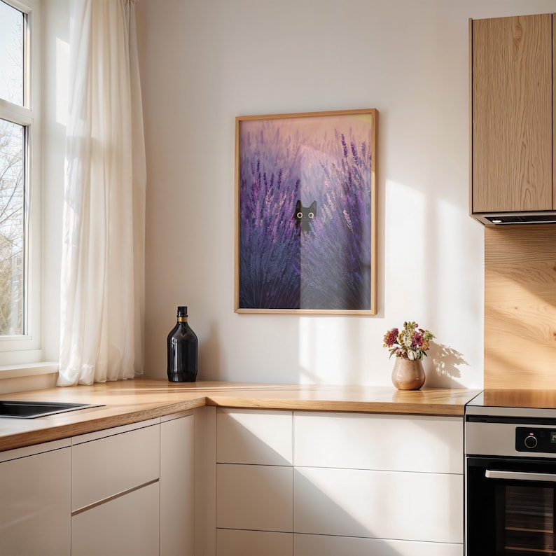 May include: A framed print depicting a black cat amongst purple lavender. The artwork is in a wooden frame and hangs in a kitchen with white cabinets and a wooden worktop. A black bottle and a vase of flowers are also visible on the worktop.