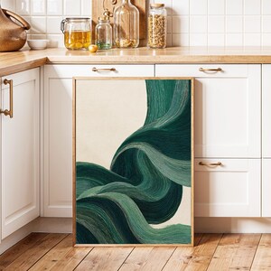 May include: A framed abstract art print featuring flowing, layered brushstrokes in shades of green and teal against a cream background. The artwork is displayed on a wooden floor, leaning against a white cabinet in a kitchen setting.