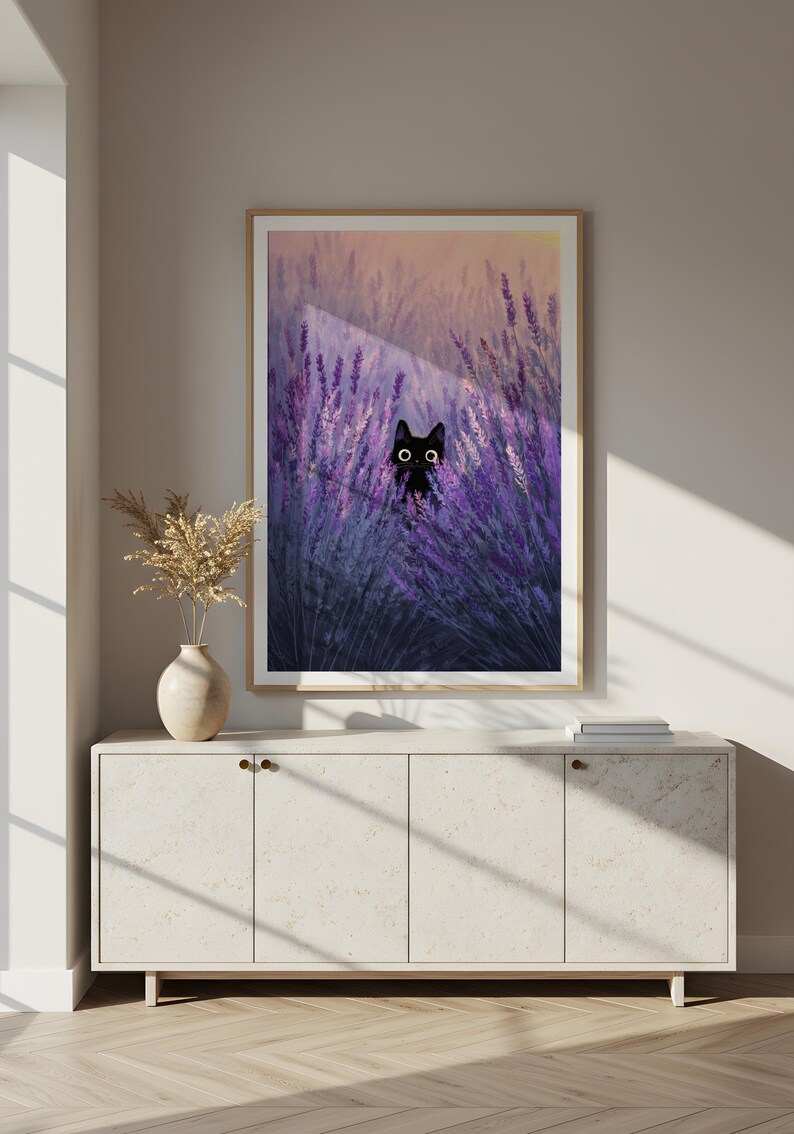 May include: A framed art print depicting a black cat amongst purple lavender. The cat has large, round eyes. The artwork is displayed above a white cabinet with a vase and dried flowers.