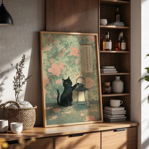 May include: A framed art print featuring a black cat interacting with a lantern. The background has a floral pattern in shades of green and pink. The print is displayed on a wooden cabinet with decorative items.