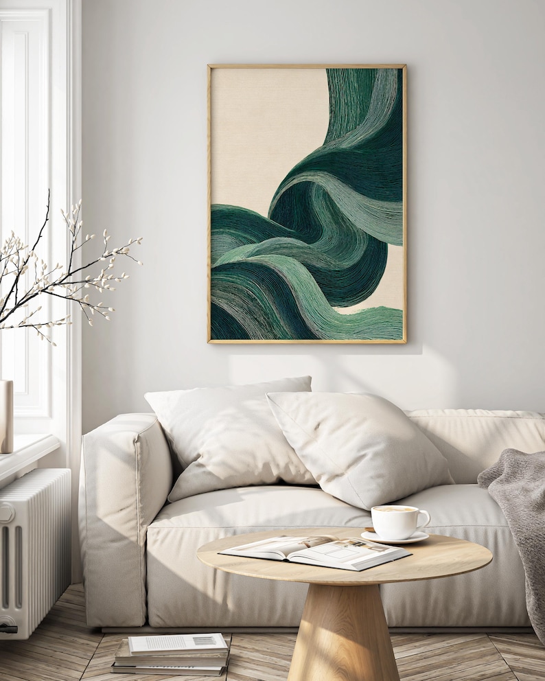 May include: Abstract art print featuring flowing, layered lines in shades of green against a cream background. The artwork is framed in a thin, gold-colored frame. The print hangs above a white sofa with pillows, a wooden coffee table, and a cup of coffee.