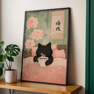 May include: Framed art print featuring a black cat in bed, holding a steaming cup of coffee. The artwork has a Japanese aesthetic, with pink peonies and a scroll with Japanese characters. The cat is on a pink comforter.