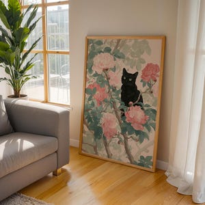 May include: A framed art print featuring a black cat perched among pink and white flowers and green leaves. The artwork is in a wooden frame and leans against a wall. The room has a sofa, a large plant, and a window with natural light.