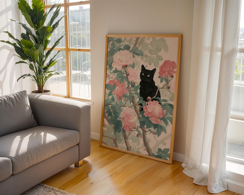 May include: A framed art print featuring a black cat perched among pink and white flowers and green leaves. The artwork is in a wooden frame and leans against a wall. The room has a sofa, a large plant, and a window with natural light.
