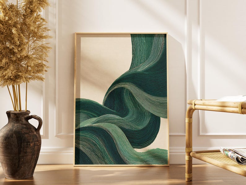 May include: Abstract art print featuring flowing, layered forms in shades of green and teal against a beige background. The artwork is framed in a gold-colored frame and leans against a white wall. A decorative vase with dried plants is on the left.