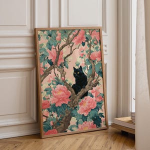 May include: A framed art print featuring a black cat perched in a flowering tree. The artwork showcases a black cat with yellow eyes amidst pink peonies and green foliage against a soft pink background. The frame is a light brown color.