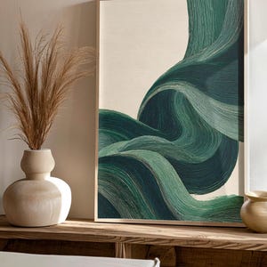 May include: Abstract art print featuring flowing, layered lines in shades of green against a cream background. The artwork is framed in a light wood frame. The image is displayed on a wooden surface, with a vase of dried plants and other decorative items.