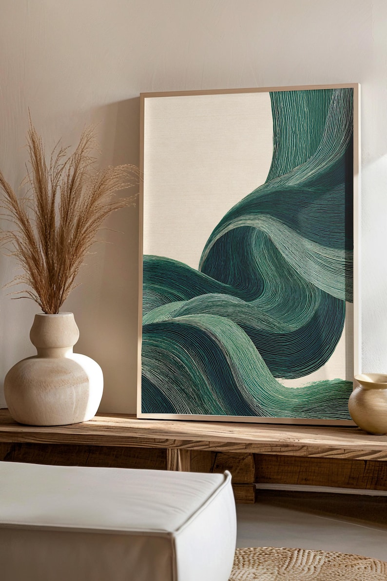 May include: Abstract art print featuring flowing, layered lines in shades of green against a cream background. The artwork is framed in a light wood frame. The image is displayed on a wooden surface, with a vase of dried plants and other decorative items.