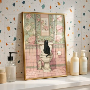 May include: A framed art print depicts a black cat sitting on a toilet in a bathroom scene. The artwork features a floral wallpaper and pink tiled walls. The frame is gold-colored, and the overall style is whimsical and artistic.