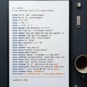 May include: A framed white poster featuring black, blue, and orange computer code text starting with # ~/.bashrc and ending with user@life:~$ followed by an orange block cursor. The text lists various bash aliases for command line operations.