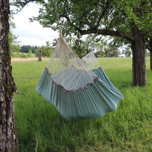 Può includere: Un'amaca sospesa, con la parte inferiore turchese e una rete color crema, è appesa a un ramo d'albero. L'amaca è ambientata su uno sfondo di erba verde e alberi, creando una scena rilassante all'aperto.