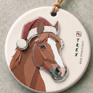 Puede incluir: Un adorno de cerámica redondo con un caballo marrón que lleva un gorro de Papá Noel rojo y blanco. El caballo tiene una mancha blanca en la cara. El adorno incluye el texto "TREX" y "CHRISTMAS 2026" en negro, con un cordón marrón.