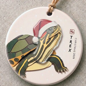 Puede incluir: Un adorno de cerámica redondo que muestra una tortuga con un gorro de Papá Noel rojo y blanco. La tortuga tiene un caparazón y piel con patrones verdes y amarillos. El texto dice "TREX" y "CHRISTMAS 2026" junto a un pequeño logotipo cuadrado.
