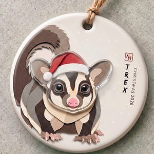 Puede incluir: Un adorno redondo de cerámica blanca con un petauro del azúcar que lleva un gorro de Papá Noel rojo y blanco. El adorno incluye un cordel marrón para colgar y el texto "TREX" y "CHRISTMAS 2026" junto a un pequeño logo cuadrado rojo.
