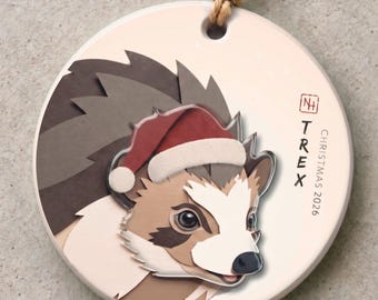 May include: A round ceramic ornament featuring a layered paper-style illustration of a hedgehog wearing a red and white Santa hat. The text reads TREX CHRISTMAS 2026 with a small square logo. A brown twine loop is attached for hanging.