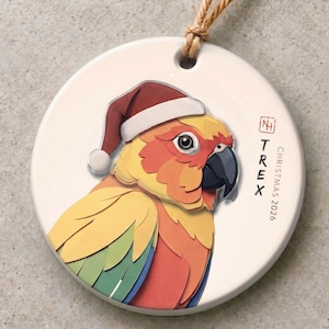 Puede incluir: Un adorno redondo de cerámica blanca con un colorido loro conure sol que lleva un gorro de Papá Noel rojo y blanco. El texto TREX y CHRISTMAS 2026 está impreso a la derecha. Un cordel marrón está sujeto en la parte superior.