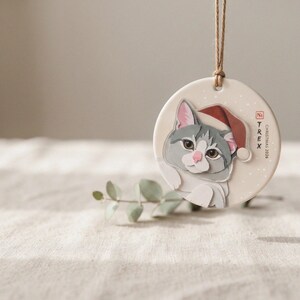 Personalized grey-and-white cat Christmas ornament hanging on an evergreen branch in a softly lit, calm holiday setting.