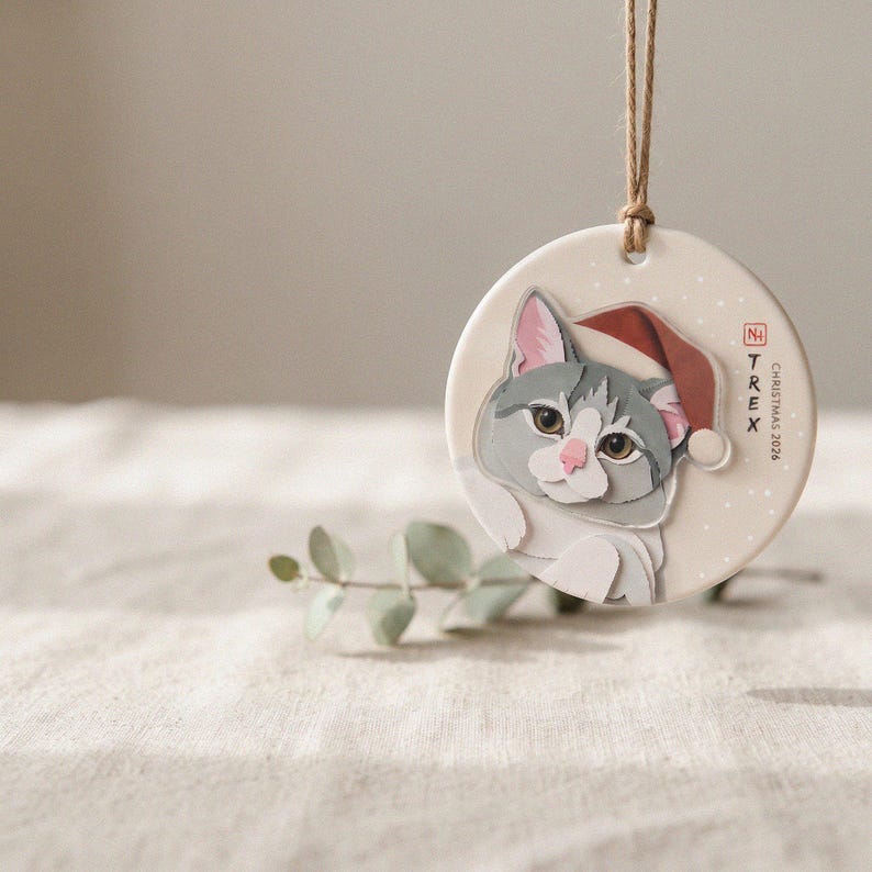 Personalized grey-and-white cat Christmas ornament hanging on an evergreen branch in a softly lit, calm holiday setting.