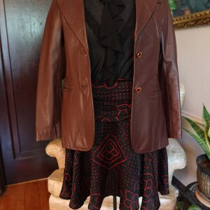 May include: A brown leather jacket with a black ruffled blouse and a black and red patterned skirt.