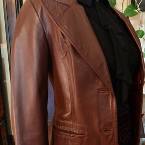May include: A brown leather jacket with a single-breasted closure and a pocket on the left side. The jacket has a collar and a long sleeve.