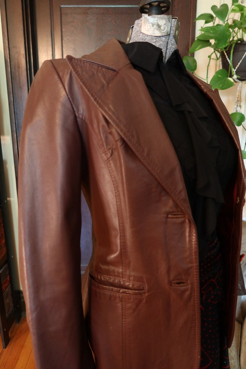 May include: A brown leather jacket with a single-breasted closure and a pocket on the left side. The jacket has a collar and a long sleeve.