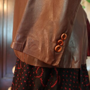 May include: A brown leather jacket with three buttons on the sleeve. The jacket is worn over a black skirt with a red pattern.