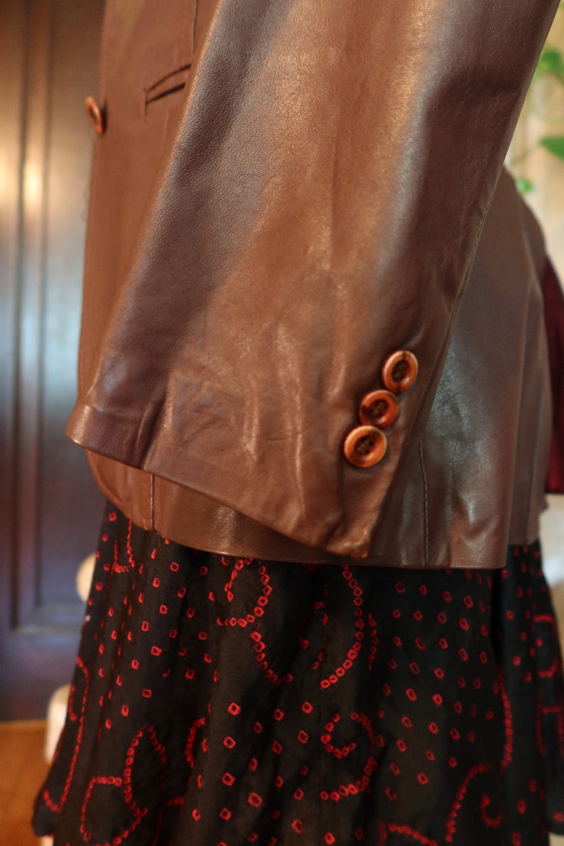 May include: A brown leather jacket with three buttons on the sleeve. The jacket is worn over a black skirt with a red pattern.