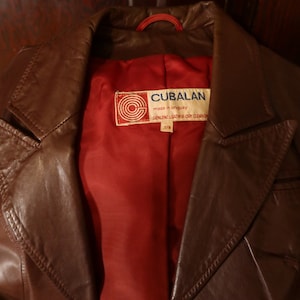 May include: A brown leather jacket with a red lining. The jacket has a label that reads "CUBALAN made in uruguay GENUINE LEATHER DRY CLEAN ONLY 7/8"