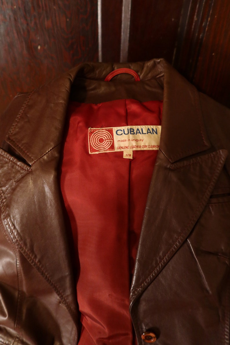 May include: A brown leather jacket with a red lining. The jacket has a label that reads "CUBALAN made in uruguay GENUINE LEATHER DRY CLEAN ONLY 7/8"