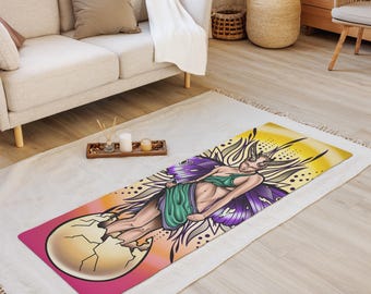 May include: A rectangular yoga mat featuring a fantasy illustration of a woman with butterfly wings emerging from a cracked egg. The design includes purple, green, and yellow hues, with a pink border. The mat is on a white rug.