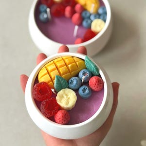 Op de afbeelding: Twee witte keramische kommen gevuld met paarse was, versierd met kaarsen in de vorm van fruit. De kaarsen lijken op mango, bosbessen, frambozen, aardbeien en bananenschijfjes. Eén kom wordt in een hand gehouden.