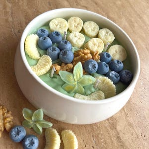 Pode incluir: Uma taça de cerâmica branca cheia de cera verde-clara, decorada com peças de cera em forma de fatias de banana, mirtilos, metades de noz, gomos de citrinos e uma pequena flor. Mirtilos e gomos de citrinos de cera adicionais estão ao lado.