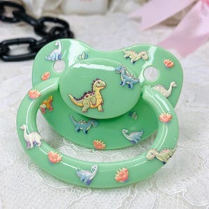 May include: A mint green pacifier featuring various cartoon dinosaurs in yellow, blue, and white. The pacifier has a rounded shield and a loop handle, with small floral accents. The dinosaurs are in different poses.