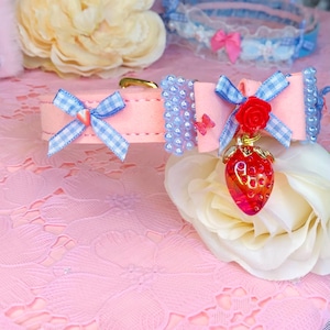 May include: Pink pet collar with blue gingham bows, light blue beaded accents, and a red strawberry charm. The collar has a gold-colored buckle and a small red rose. The background includes a pink floral lace.