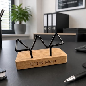 Könnte beinhalten: Ein Schreibtisch-Accessoire mit einem rechteckigen, hellbraunen Holzsockel, in den der Text EPHK Maier eingraviert ist. Auf der Oberseite des Holzsockels befindet sich ein schwarzer Metalldrahtrahmen in einer geometrischen, zackigen Form.