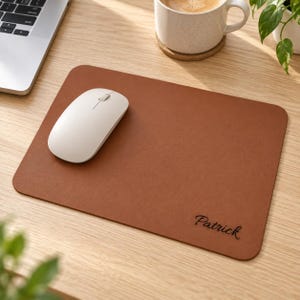 Peut inclure: Un tapis de souris rectangulaire en cuir marron aux coins arrondis, portant le prénom Patrick gravé en écriture cursive noire dans le coin inférieur droit. Une souris d'ordinateur sans fil blanche repose sur le côté gauche du tapis.
