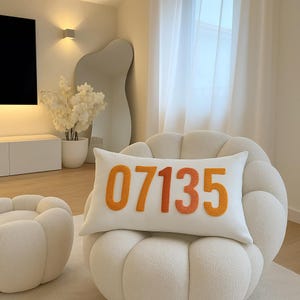 Op de afbeelding: Een wit rechthoekig kussen met het nummer "07135" in oranje en roodoranje. Het kussen ligt op een crèmekleurige, afgeronde fauteuil. De kamer heeft een neutraal kleurenpalet met een moderne esthetiek.