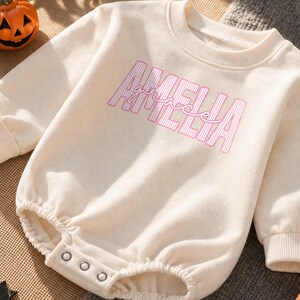 May include: A cream-colored long-sleeve baby romper featuring the name AMELIA in pink striped block letters with the word grace written in a cursive pink script overlay. The garment has a ribbed crew neckline, cuffs, and a snap-button closure.