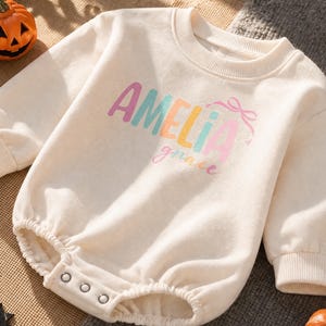 May include: A cream-colored long-sleeve baby romper with ribbed cuffs and a crew neckline. The front features the name AMELIA in multi-colored block letters, a small pink bow graphic, and the word grace in pink cursive script text.